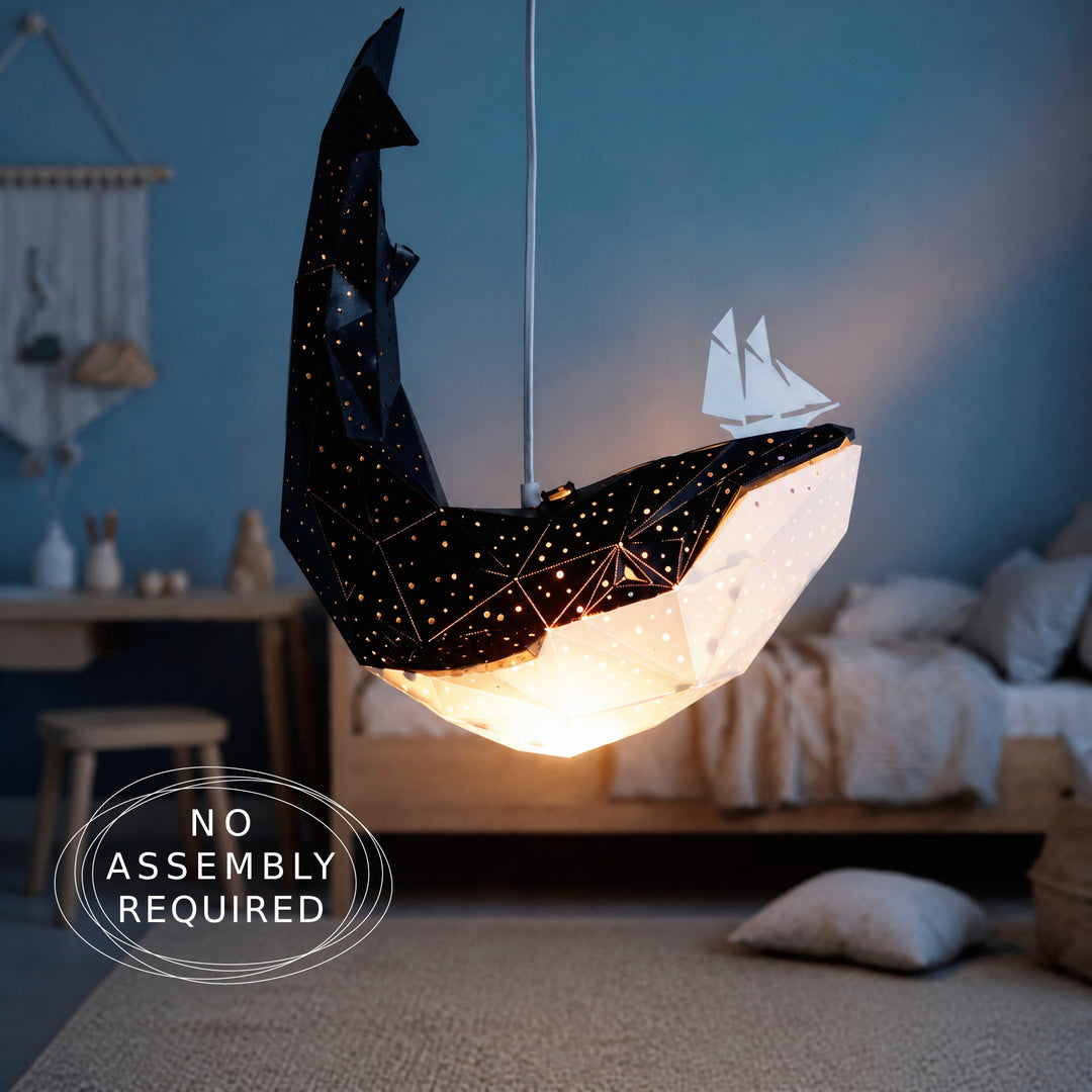A whale-shaped hanging pendant light with a faceted geometric design, shown illuminated in a bedroom setting. The top half is black with small gold star-like dots, and the bottom half glows warm white.