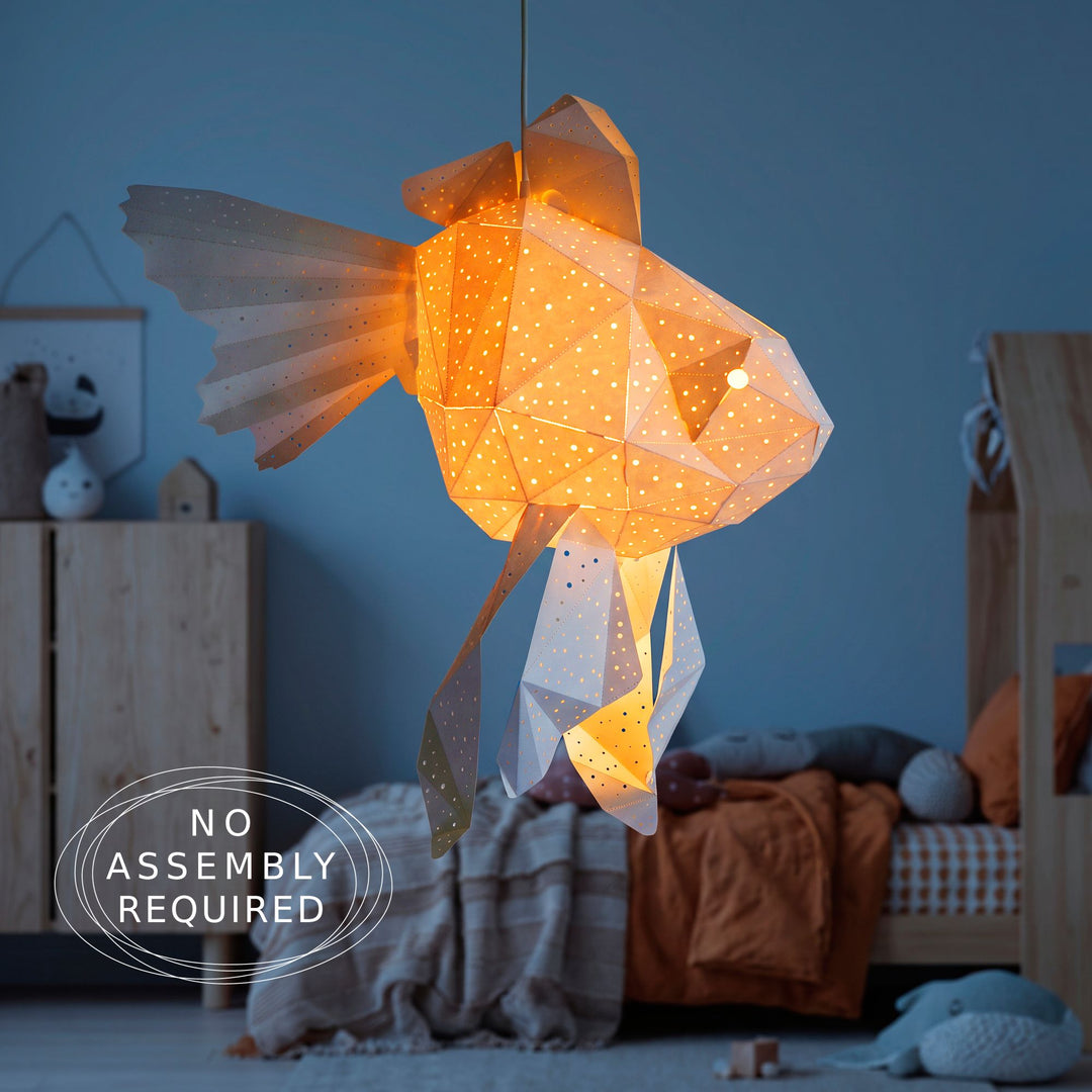 An orange and white paper goldfish pendant light glowing above a child's bed against a deep blue wall, with a no assembly required badge in the corner.