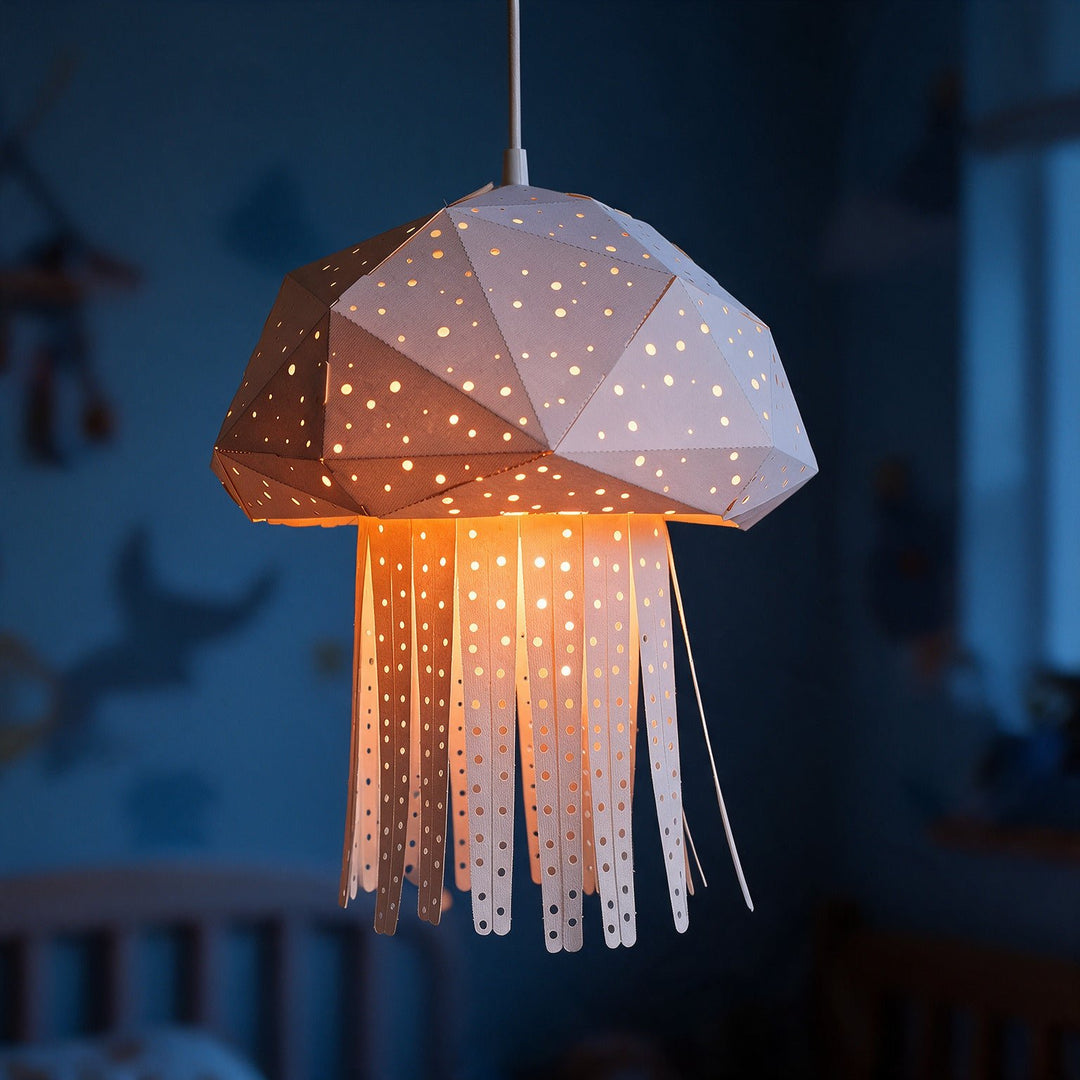 A jellyfish-shaped hanging pendant lamp made from white paper with cutout star patterns, glowing warm yellow-orange from an internal light, shown hanging in a dimly lit blue bedroom.