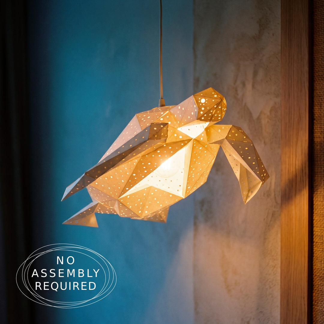 A geometric turtle-shaped pendant light made of translucent paper with cutouts, glowing warm yellow from within. It is shown hanging against a split blue and textured beige wall, with a 'NO ASSEMBLY REQUIRED' badge in the bottom-left corner.