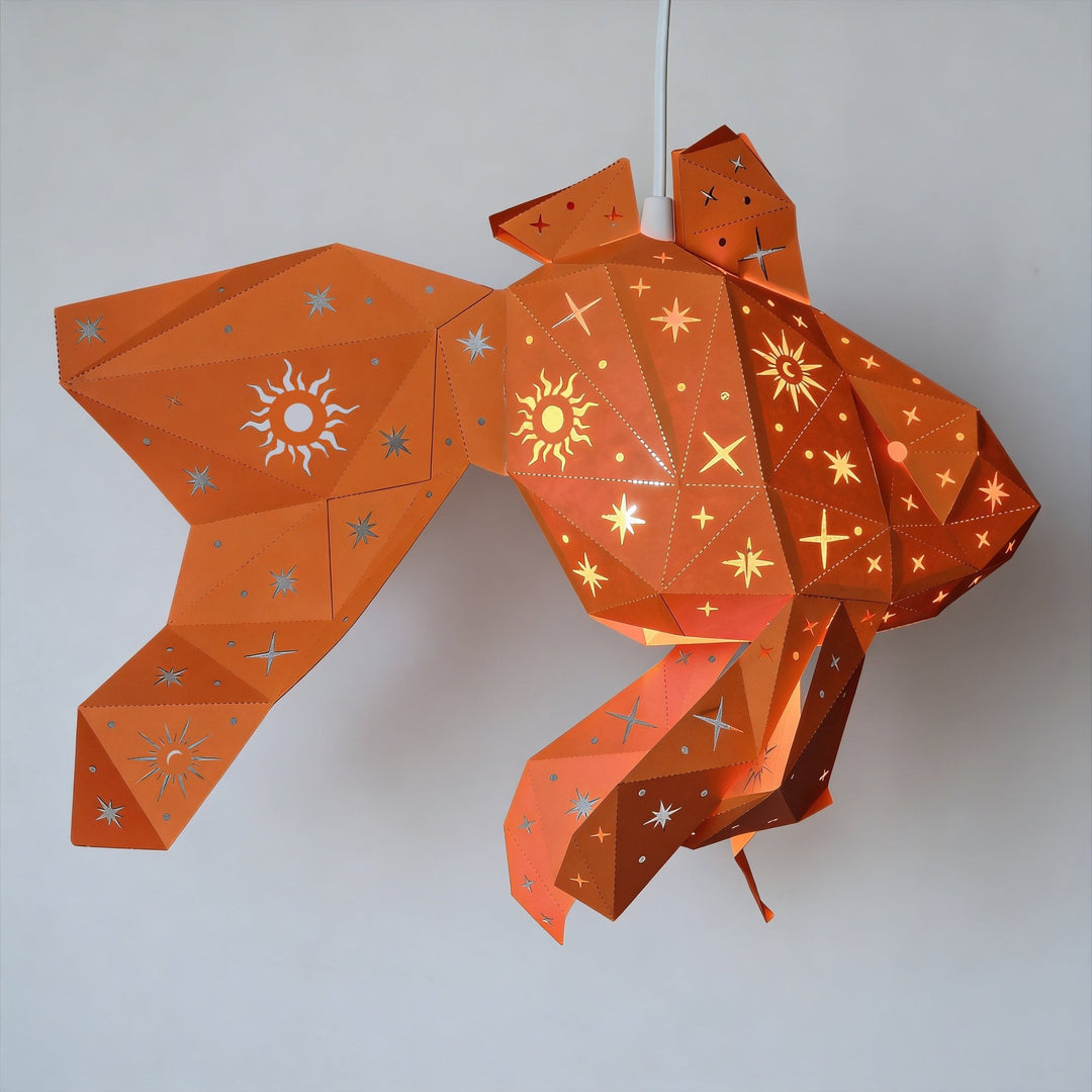 An orange paper lantern shaped like a fish, decorated with silver and gold star and sun patterns, hanging against a plain light gray wall.