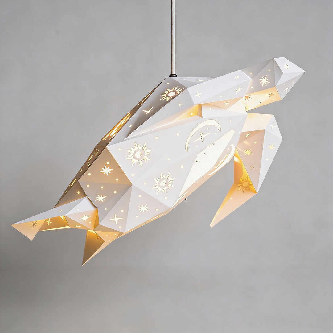 A geometric paper lantern-style pendant light shaped like a sea turtle, suspended from a thin rod against a plain light gray wall. The lantern is white with gold star and moon cutouts.