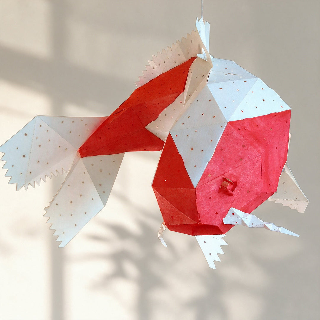 A geometric, faceted fish ornament made from red and white patterned paper, suspended against a warm beige wall with soft shadows.
