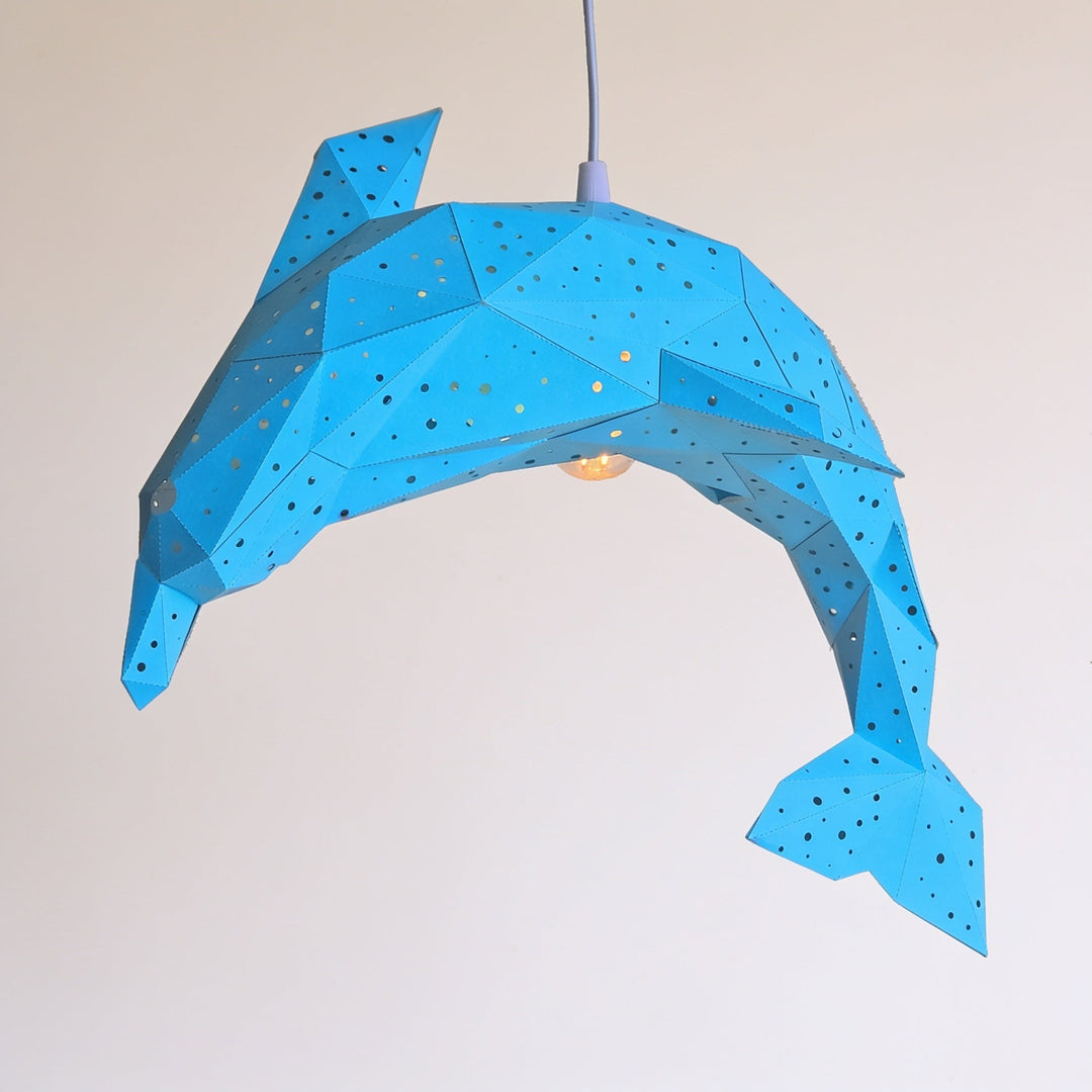 Suspended from a cord against a plain background, the Vasili Lights Bubble Dolphin Lantern – DIY Paper Night Light Kit features a blue geometric dolphin shape adorned with small holes, creating a charming and calming ambient glow—a perfect night light for any space.