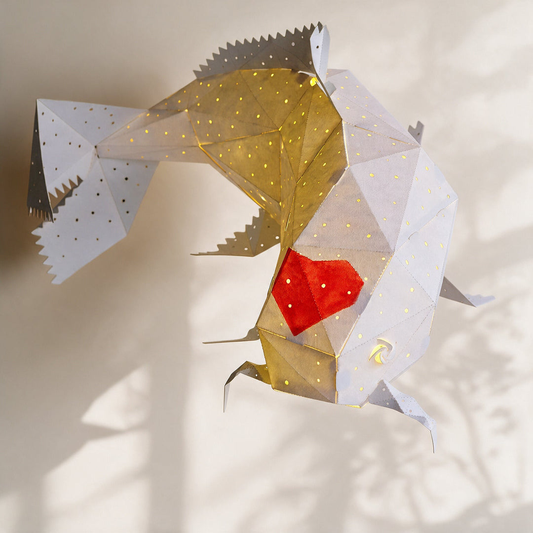 A geometric fish-shaped hanging lampshade made of white paper with small perforations, a red heart detail, shown against a softly lit beige wall with tree shadows.