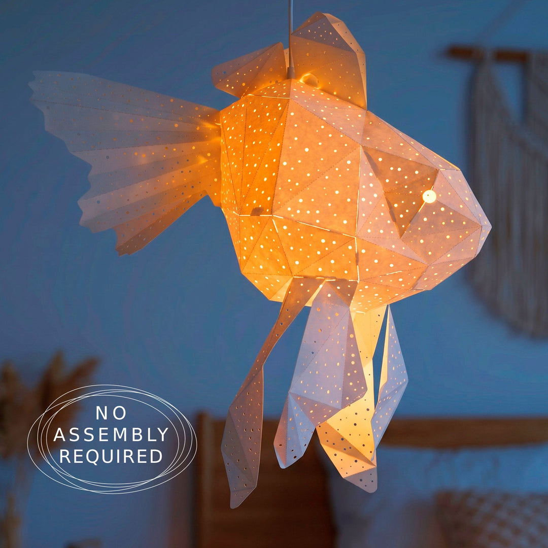 A geometric origami-style fish pendant light glowing warm white with many small cutout dots, hanging in a dimly lit bedroom. 