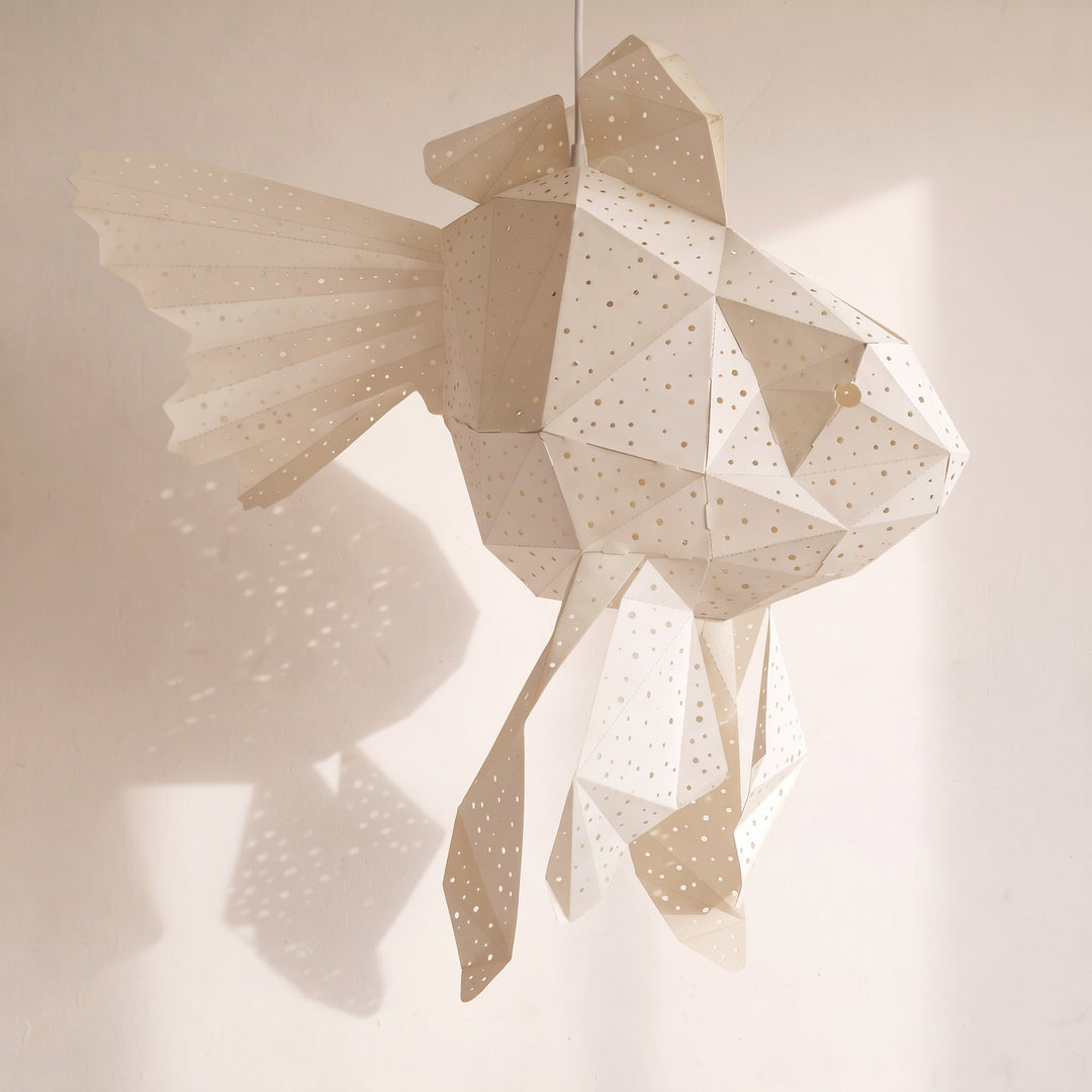 A geometric fish-shaped hanging light fixture made of white paper with small star-like dots, suspended against a warm beige wall that casts soft shadow patterns.