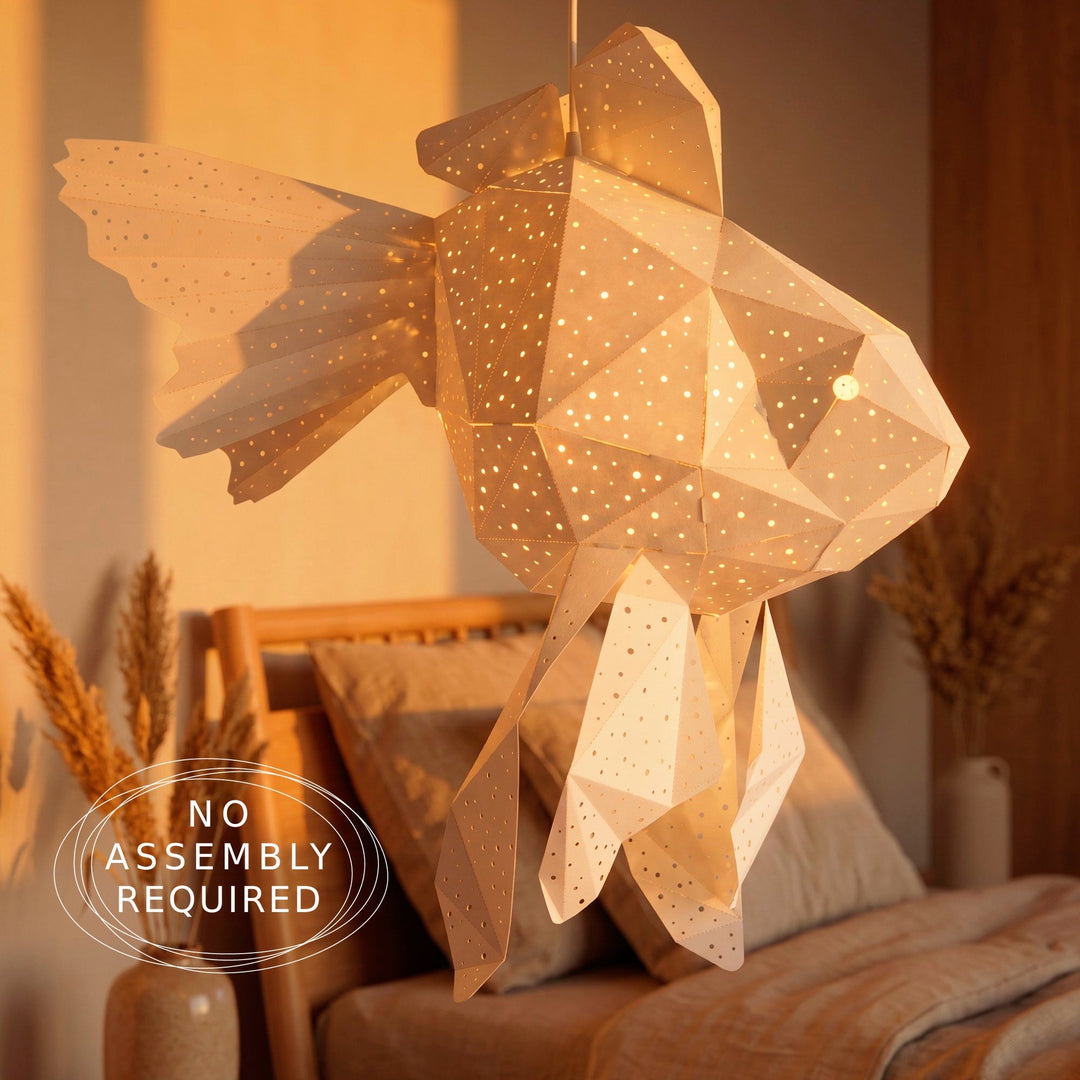 A geometric fish-shaped hanging lamp made of translucent paper with many small cutout holes, glowing warm yellow in a bedroom setting. A bed, pillows, and dried decorative plants are visible in the background.