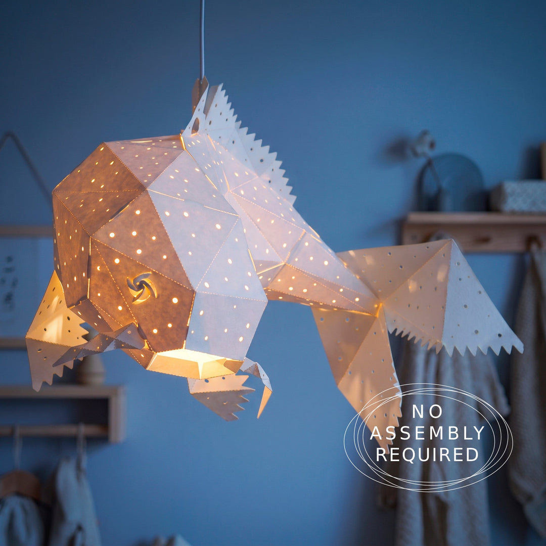 A geometric fish-shaped pendant light made of white paper with laser-cut star and wave patterns, glowing with warm yellow light from within. The light is shown hanging against a blue wall in a styled room setting.