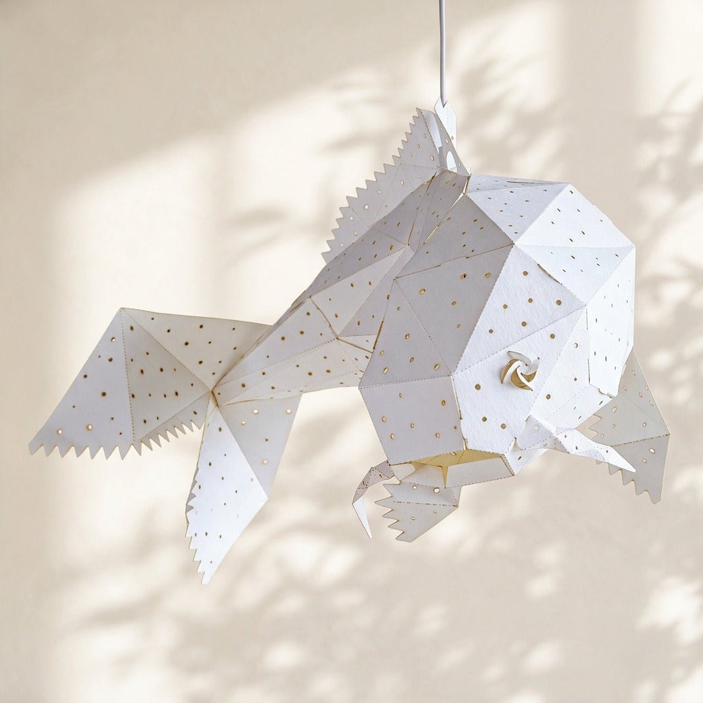 A white geometric fish-shaped hanging lamp made from folded paper, with small dot cutouts, shown against a softly lit beige wall with dappled shadows.