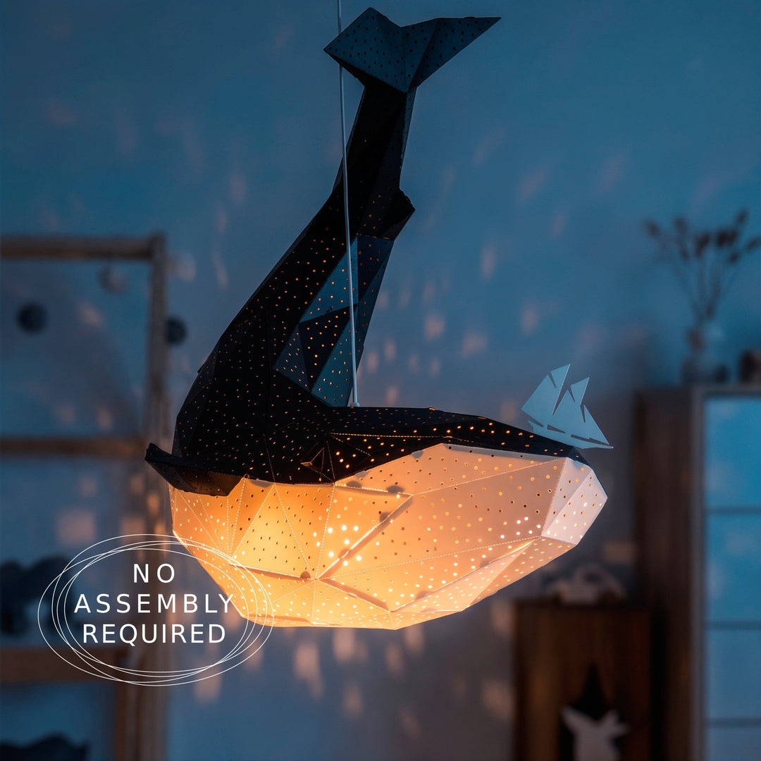 A whale-shaped hanging pendant light with a warm glowing interior and star-like cutouts, shown in a dimly lit room with a small sailboat figure beside it.