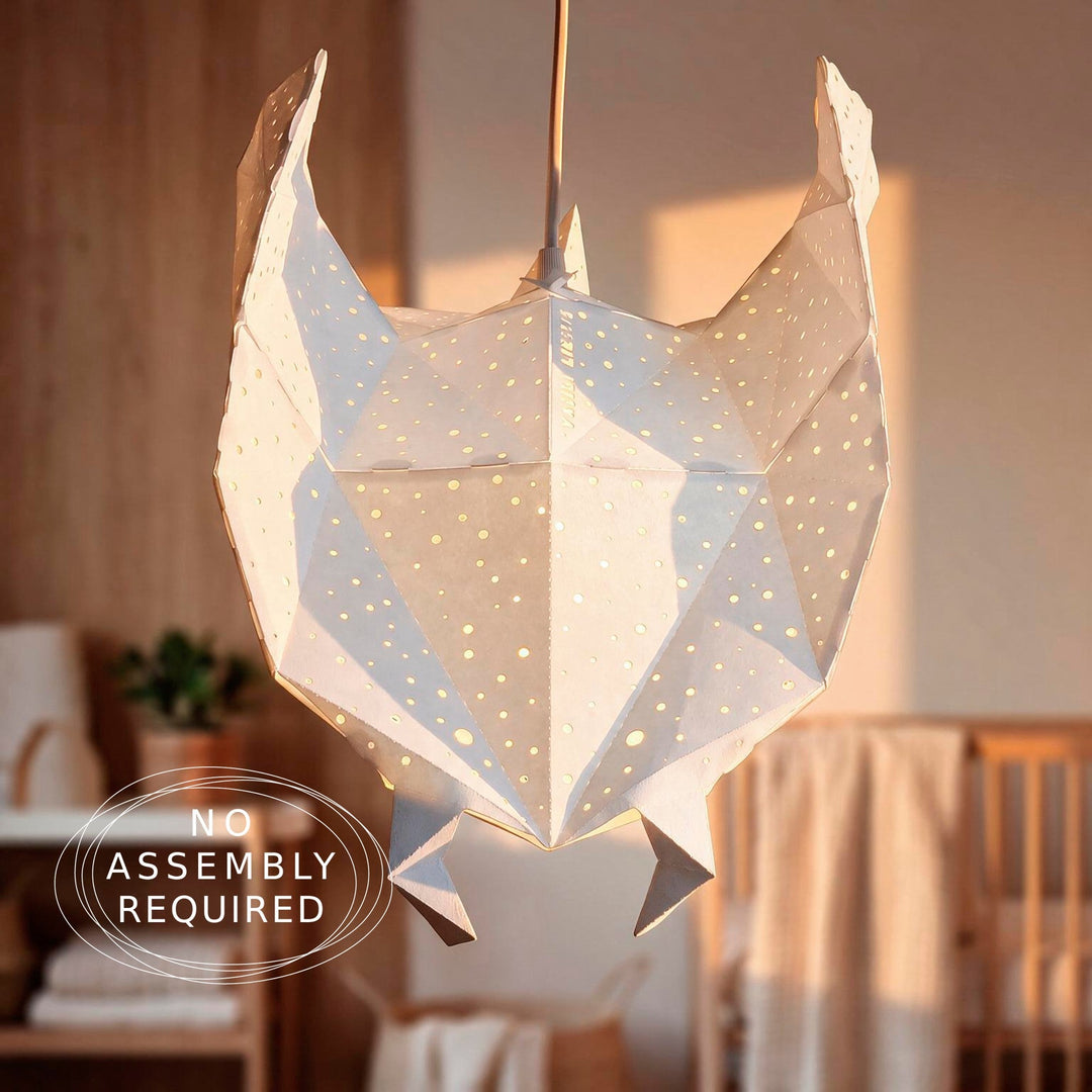 A faceted, geometric hanging pendant light made from white paper with small cutout star-like holes, shown illuminated in a softly lit nursery or bedroom setting.