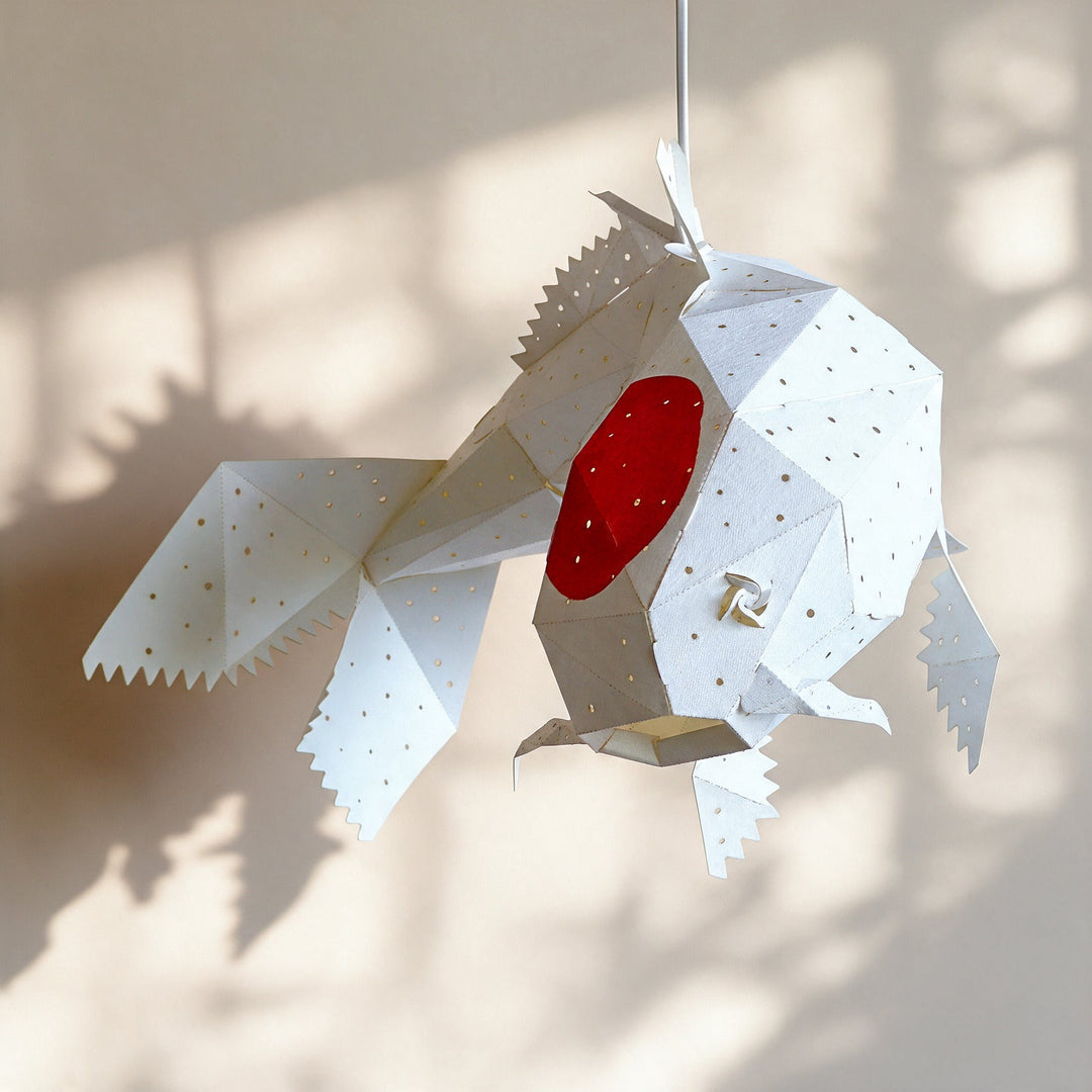 A white geometric fish-shaped hanging lamp with a red oval center, photographed against a warm beige wall with soft shadow patterns.