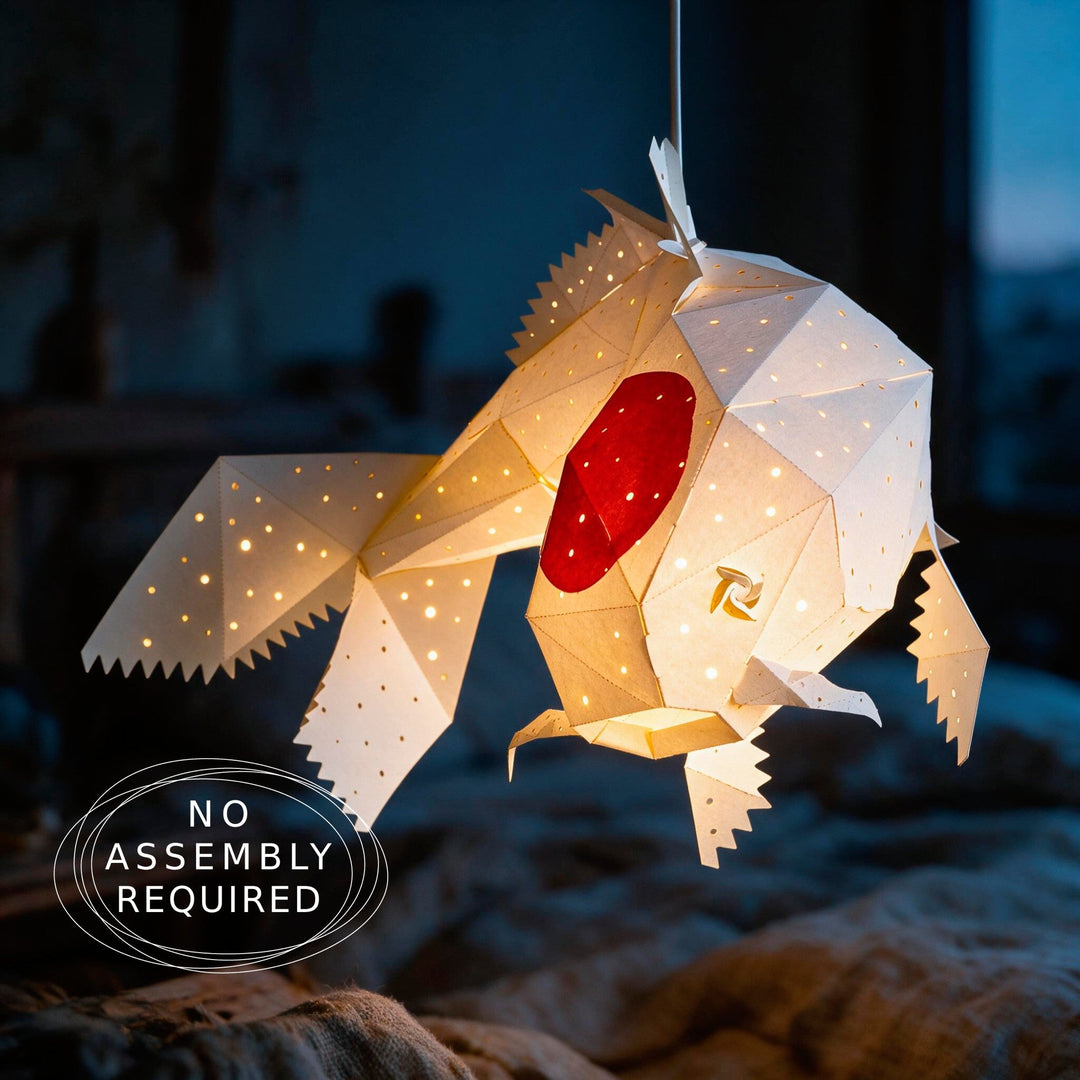 A geometric origami fish hanging lamp glowing with warm light, featuring a red circular eye and small gold details, shown against a dark blurred background.
