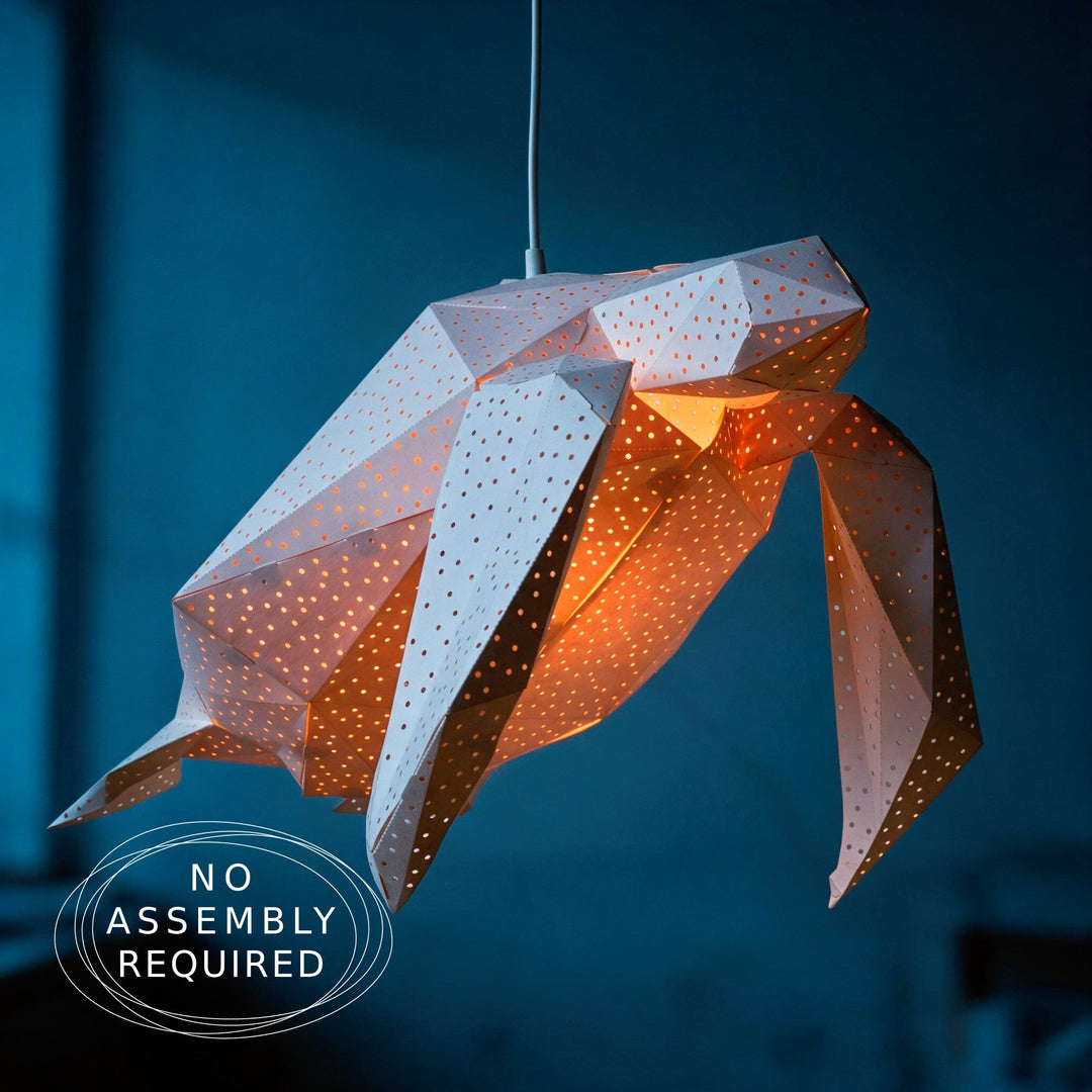 A geometric paper pendant light shaped like a stylized sea turtle, suspended by a thin cord against a dark blue background. The lantern has white outer panels with small perforations and warm orange light glowing from within.