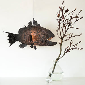 A black fish-shaped Nautical Ceiling Lamp hanging above a table next to a vase with a dark branch of a bush or tree in it.