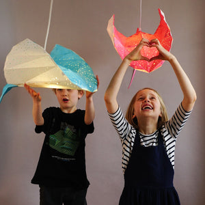 Two children are holding up Ray Origami Lamp lanterns inspired by manta rays, radiating a calming light.